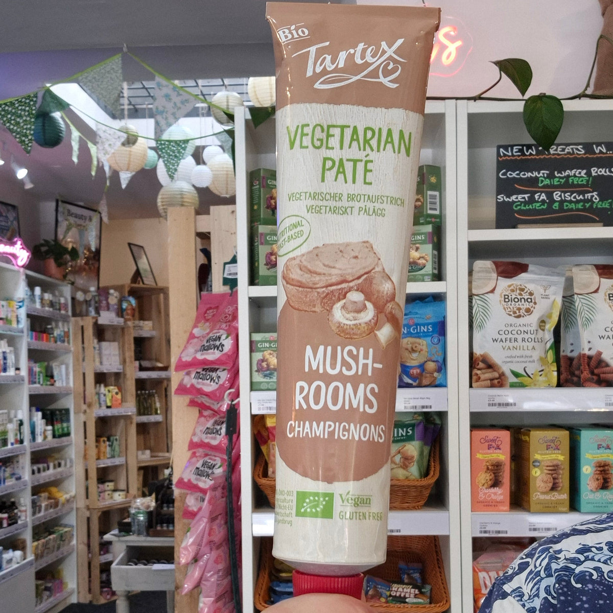A hand holding a tube of Tartex mushroom pate in a retail store setting with shelves of products in the background.