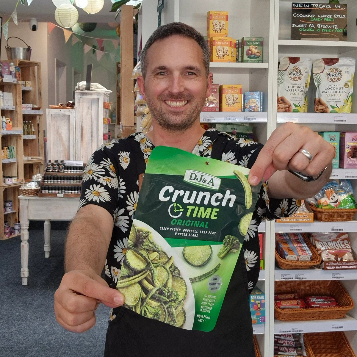 A man in a black floral shirt and black apron holds up a bag of D&G A Crunch + Time snack mix in a retail shop setting.