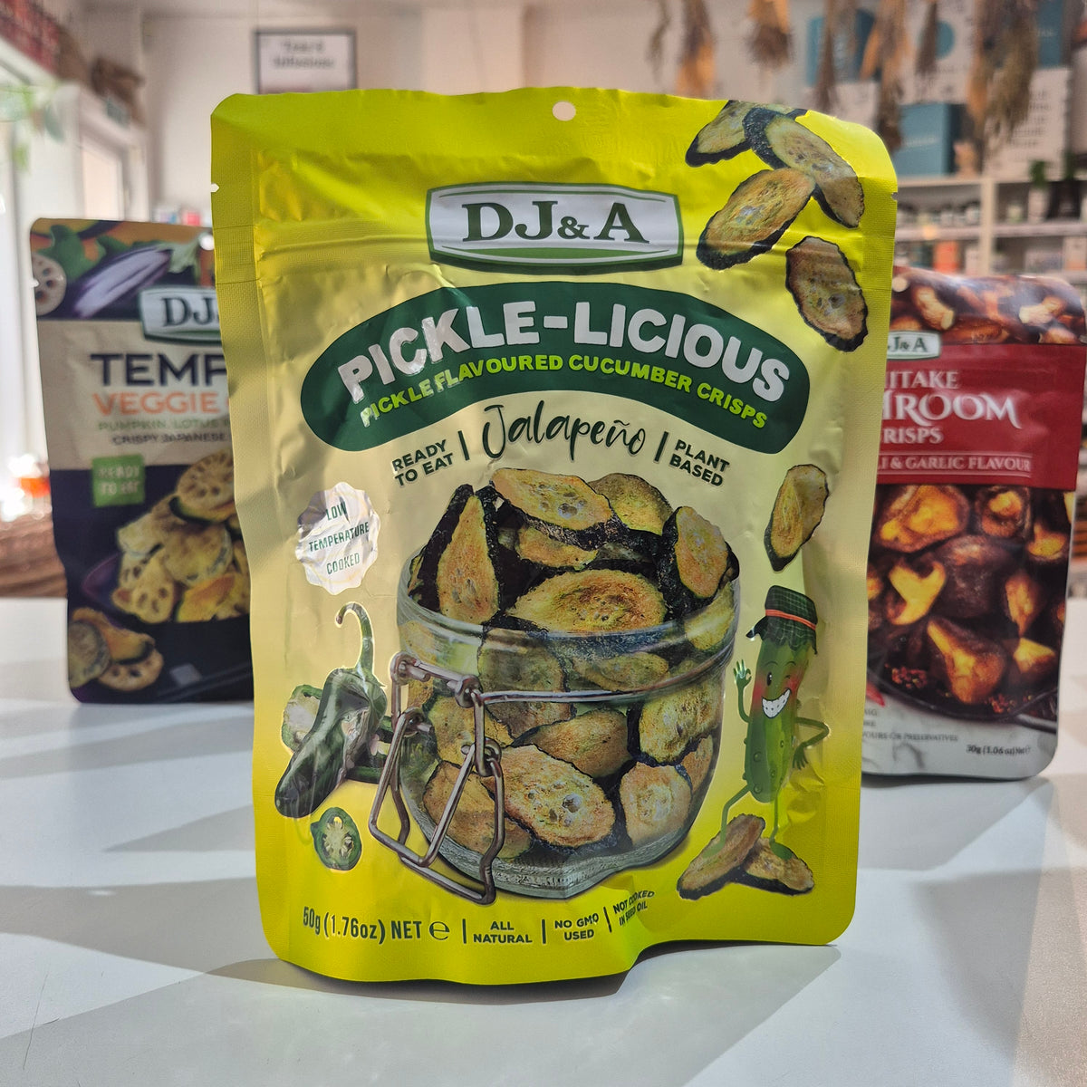 A bag of DJ&A Pickle-Licious Jalapeño pickle cucumber crisps in a bright yellow stand-up pouch, displayed on a countertop with other DJ&A snack packages blurred in the background.