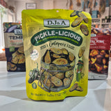 A bag of DJ&A Pickle-Licious Jalapeño pickle cucumber crisps in a bright yellow stand-up pouch, displayed on a countertop with other DJ&A snack packages blurred in the background.