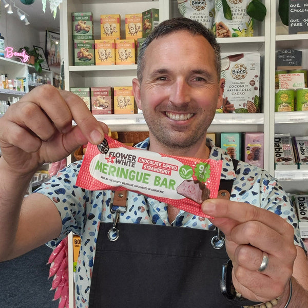 A smiling man in a patterned shirt and black apron holds up a packaged chocolate bar in front of a store shelf filled with other products. The package reads 'Flower White Chocolate Strawberry Meringue Bar'.