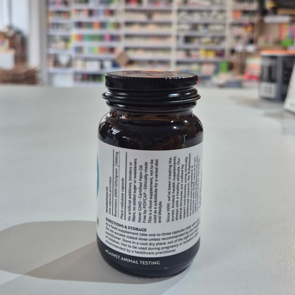 Bottle of Viridian Bromelain 500mg on a counter in a store setting. Brown supplement bottle with label on a light surface, blurred store shelves in the background