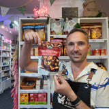 A man in a cafe or shop setting holds up a bag of Mushroom Crisps The background features shelves stocked with various packaged snacks and treats, with a pink neon 'Treats' sign hanging above.
