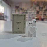 A small white ceramic ghost figurine holding a heart stands next to a square kraft paper tag printed with the words 'Bestie Boos' on a blurred indoor background.