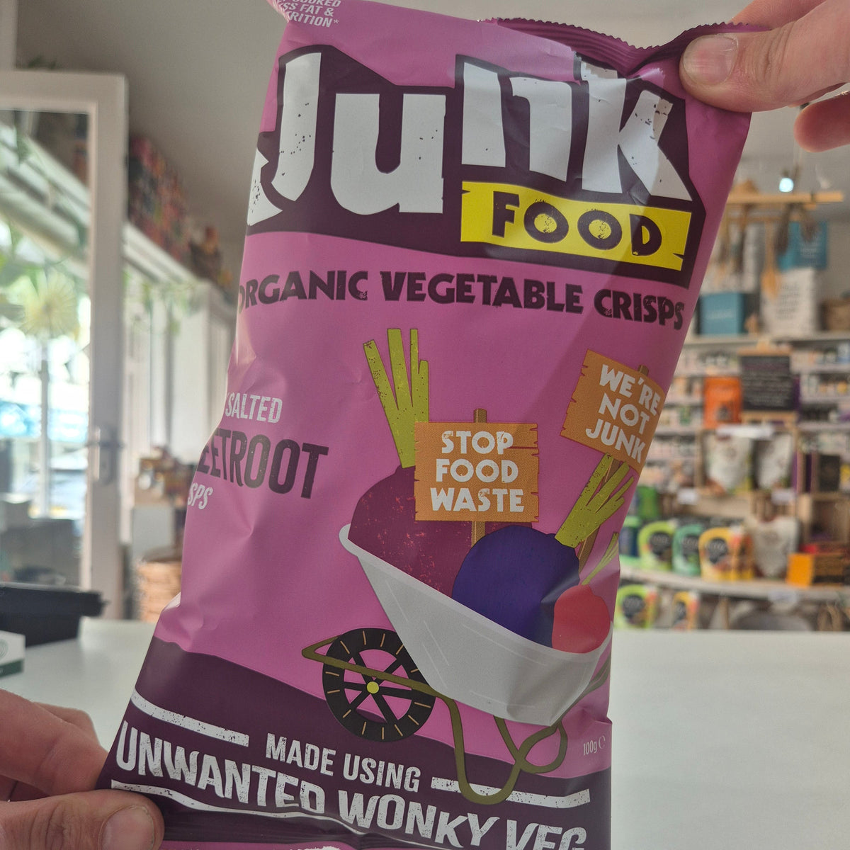 Purple package of 'Junk Food' organic vegetable crisps held by a hand in a store setting.