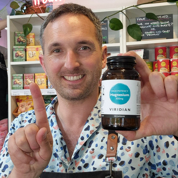 A smiling man in a patterned shirt holds up a dark amber bottle of VIRIDIAN High Potency Magnesium 300mg. The background shows a white shelving unit filled with various colorful supplement boxes and jars.
