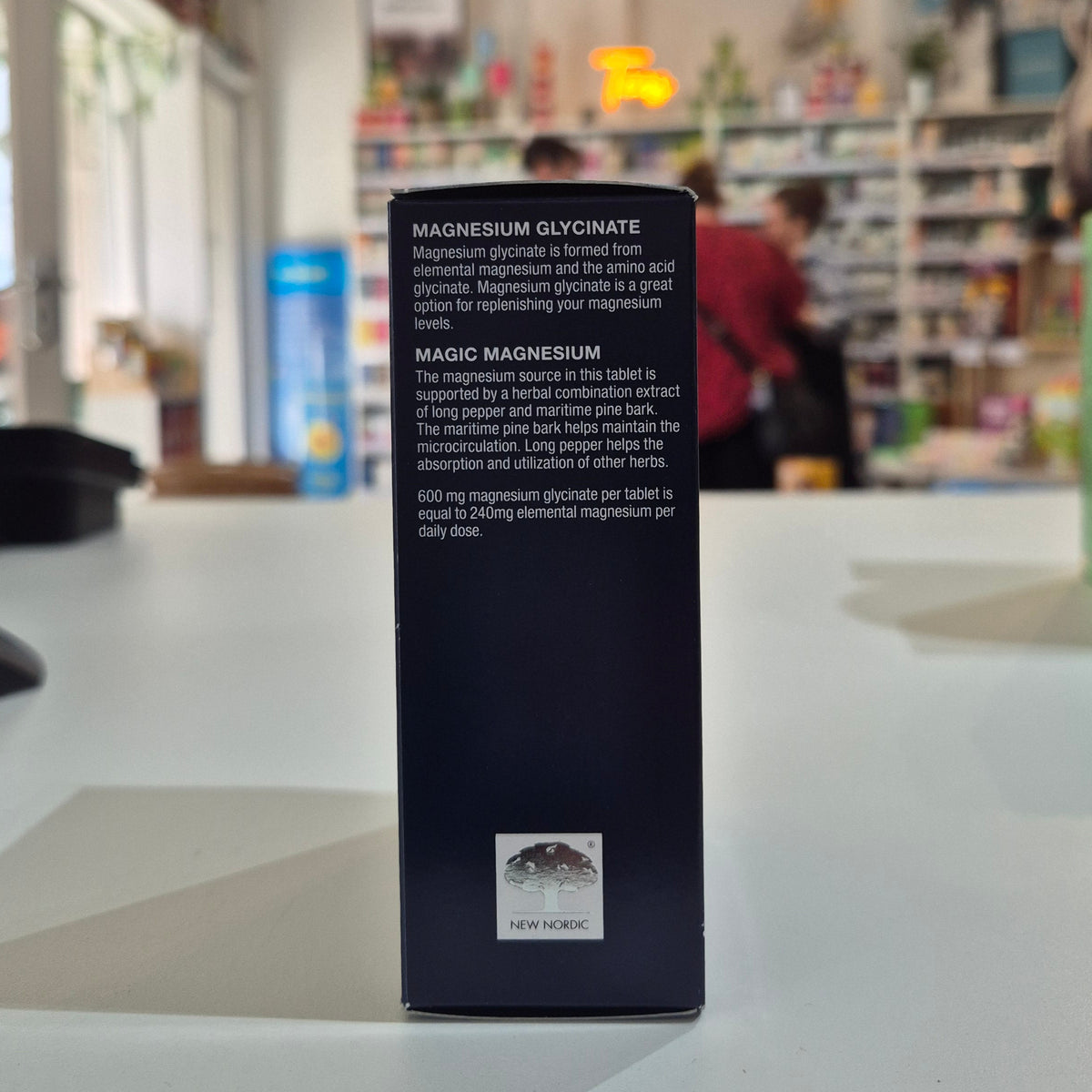 Box of New Nordic Magic Magnesium Glycinate 600 mg supplement on a blurred store shelf background. Black supplement bottle label with text on a blurred store background