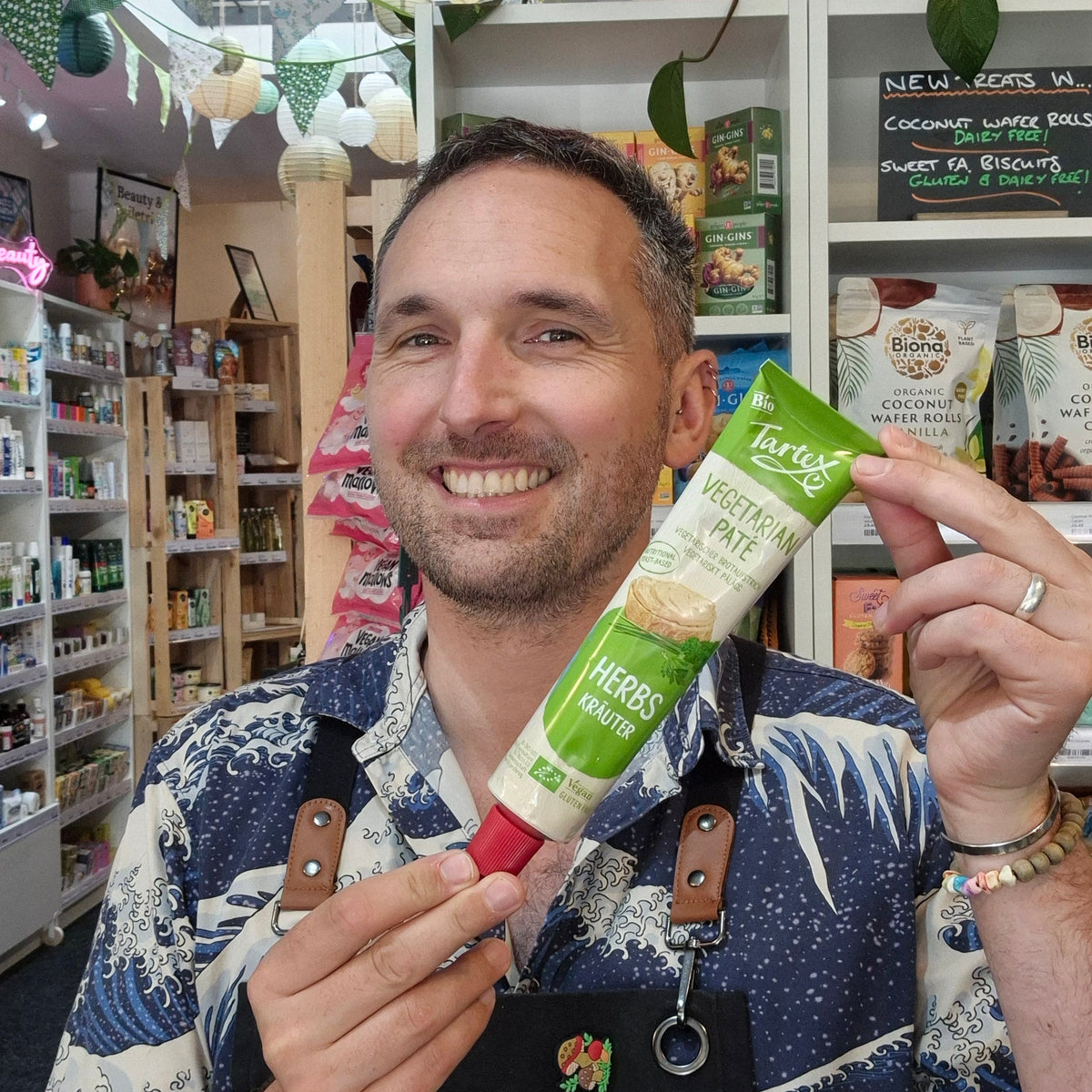 A smiling man in a black and blue wave-patterned shirt holds a green and white tube of 'Health & Remedy' Tartex supplement in front of a store shelf.