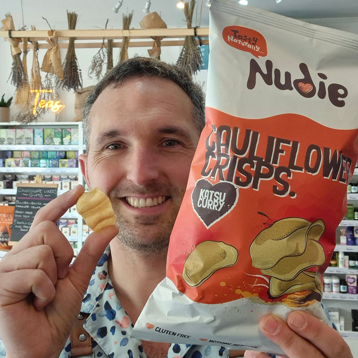 Person holding a Nudie Potato Crisps bag with a store interior in the background