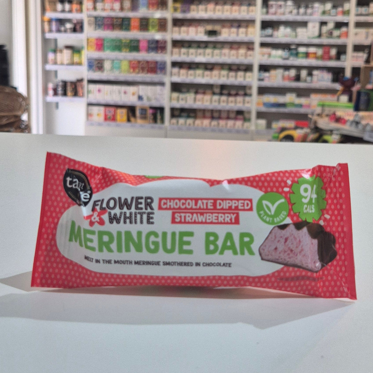 A packaged meringue bar in a red and white wrapper labeled 'Flower & White' and 'Chocolate Dipped Strawberry', placed on a white surface with a blurred store shelf background.