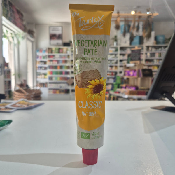 A tube of Tartex Vegetarian Pate Classic spread, featuring a yellow and white label with a sunflower graphic, lying on a white surface with a blurred store background.