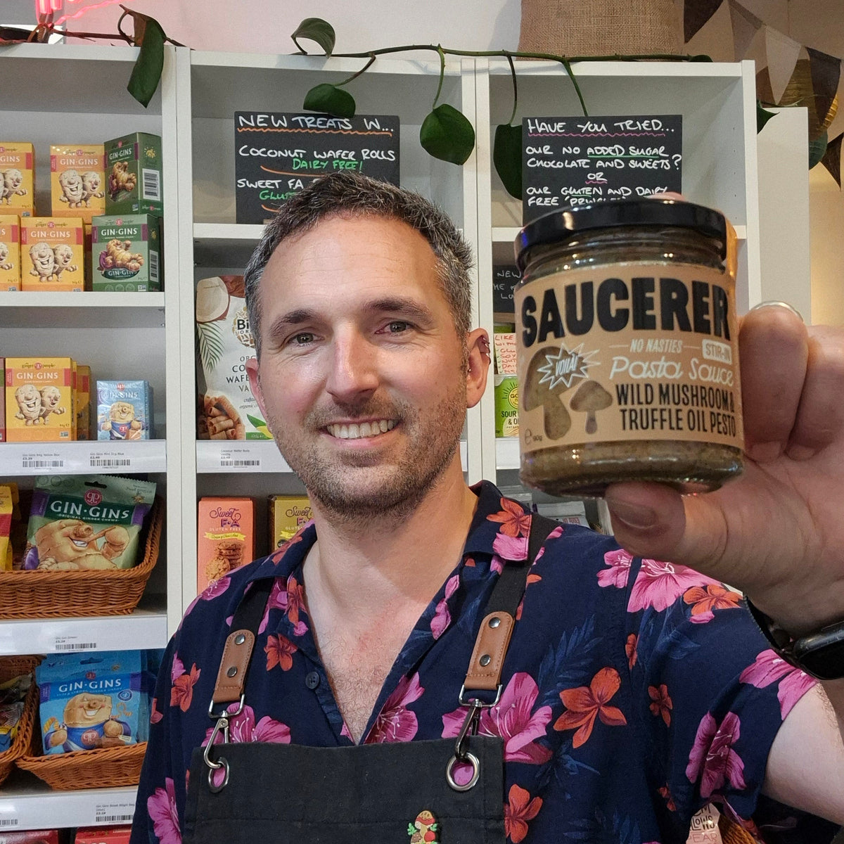 A man wearing a Health & Remedy apron holds up a jar of 'Saucerer' wild mushroom truffle oil in a shop setting with shelves of products behind him.