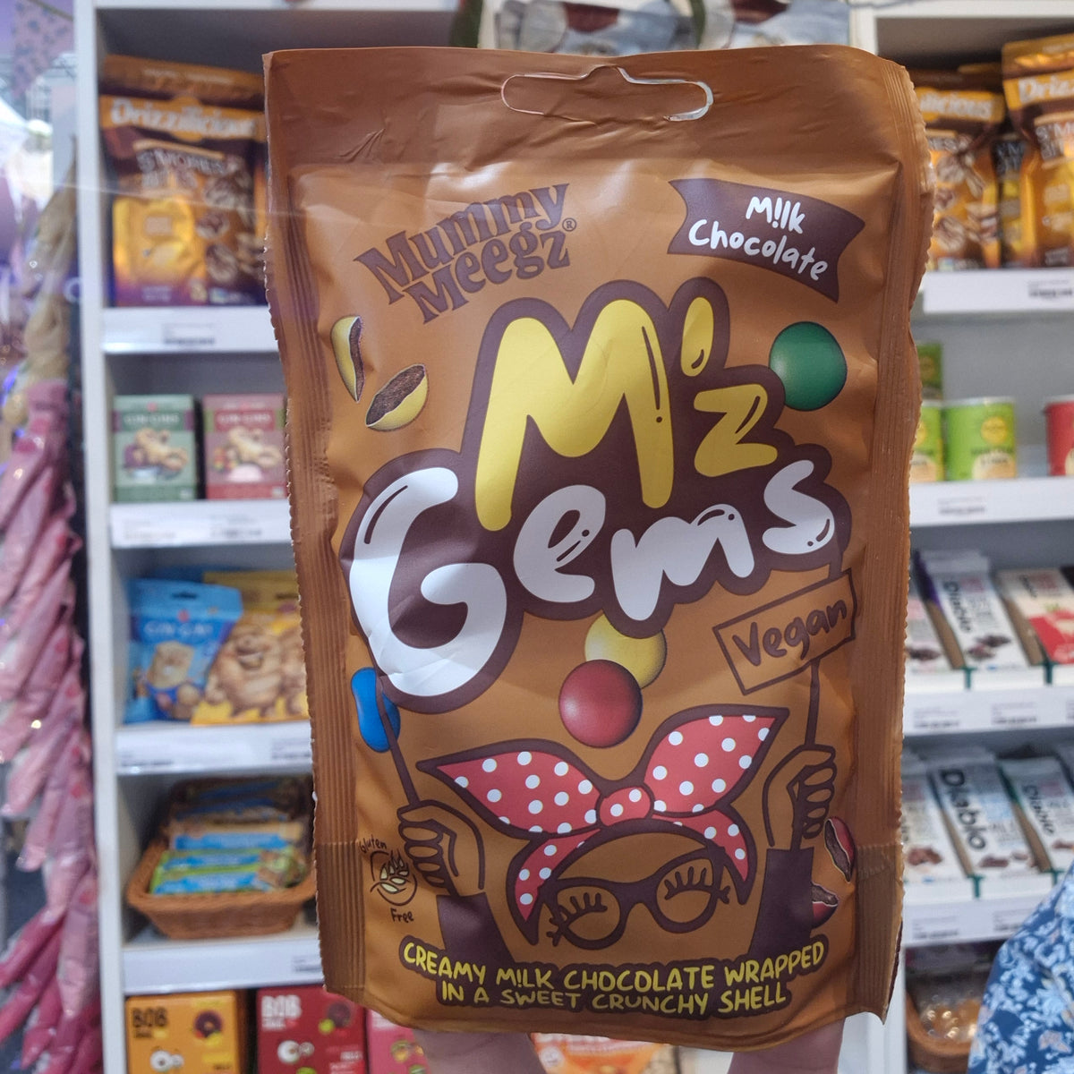 A bag of Mummy Meegz Mz Gems Vegan Milk Chocolate wrapped in a sweet crunchy shell, held up in front of a store shelf.