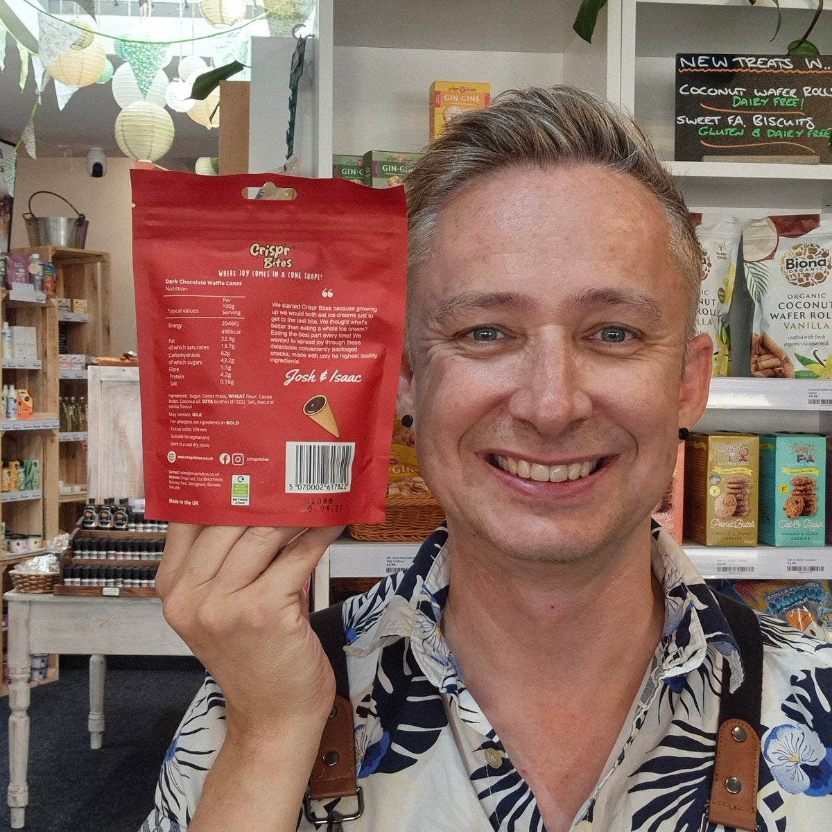 A smiling man in a patterned shirt and black apron holds up a red bag of Crispr Bites Dark Chocolate Filled Waffle Cones in a brightly lit shop shelves and a pink neon sign in the background.