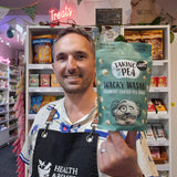 A smiling man in a patterned shirt and black apron holds up a bag of 'Taking the Pea' Wacky Wasabi Crunchy Coated Pea Snacks in a retail shop setting.