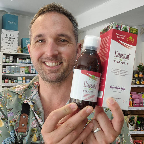 A smiling man in a floral shirt holds up a bottle of Herbetom Transit alongside its red and white product box inside what appears to be a supplement store.