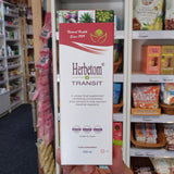 A hand holds a white and red packet of Herbetom Transit Supplement in front of a retail store shelf filled with various products.