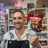 A man in a shop holding a bag of DJ&A Shiitake Mushroom Crisps (Mixed Chilli & Garlic Flavour) in front of shelves of other products.