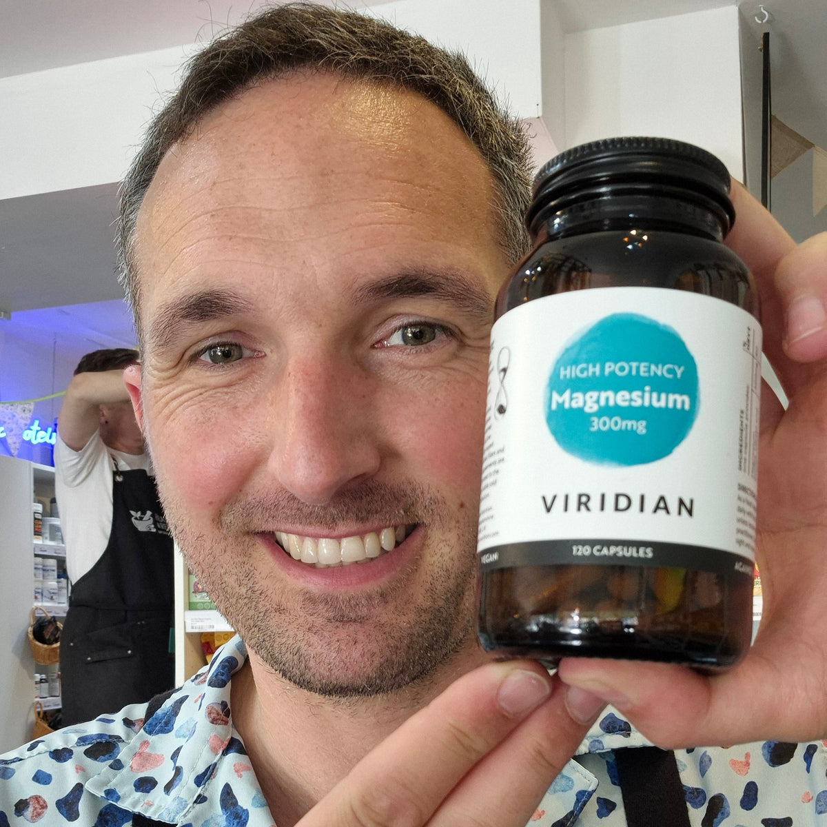 A smiling man holds up a bottle of Viridian High Potency Magnesium 300mg capsules in a brightly lit indoor setting.