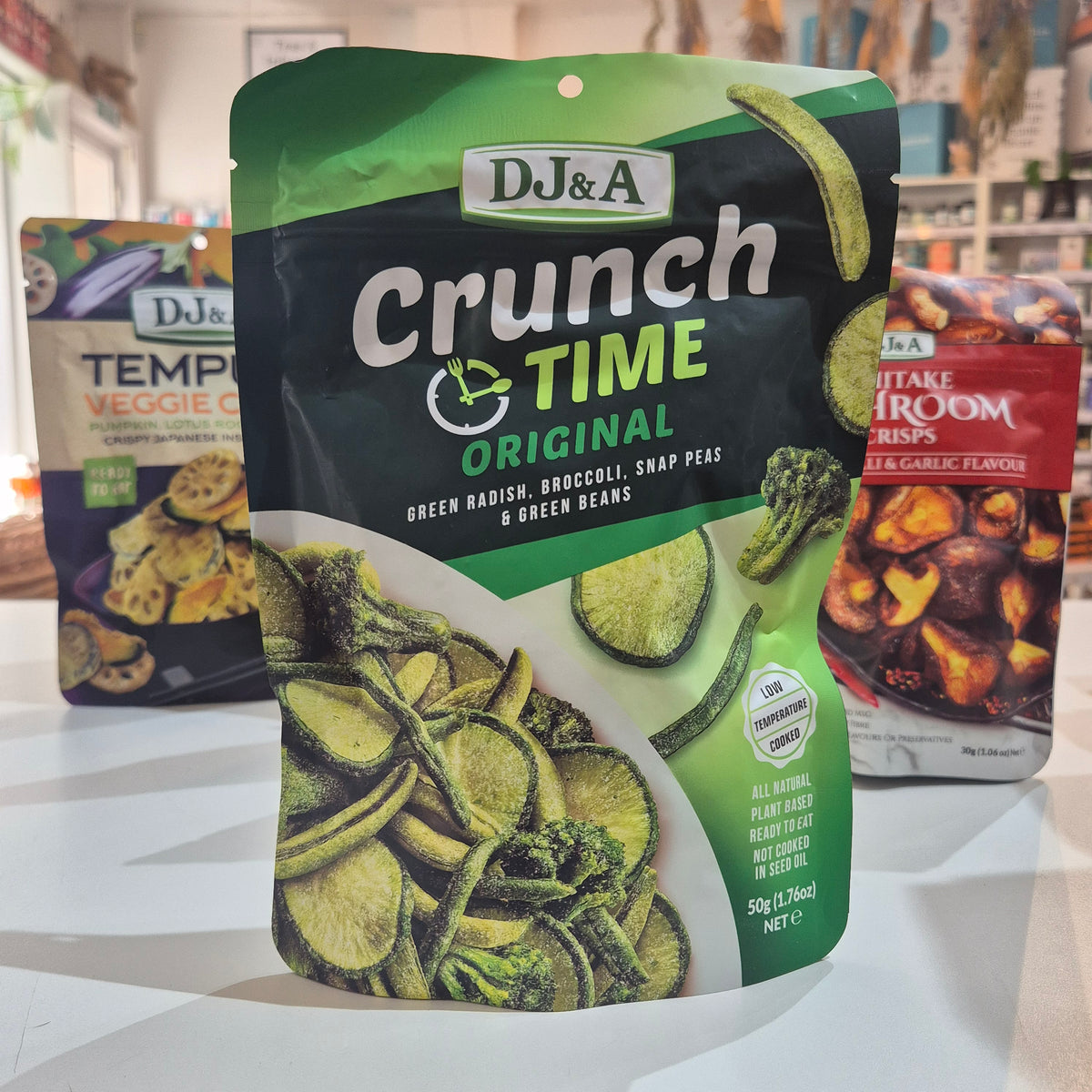 A 50g bag of DJ&A Crunch Time Original green harish, broccoli, snap peas, and green beans chips, displayed on a white surface with other snack bags in the background.