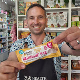 A smiling man in a shop or cafe setting holds up a Flower & White Caramel Gold Meringue Bar. The man is wearing a blue and white patterned shirt and a black apron with the Health & Remedy logo.