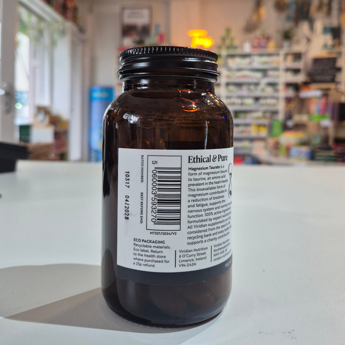 Bottle of Viridian Magnesium Taurate on a counter in a store setting. Brown glass bottle with a label on a white surface, blurred store interior in the background