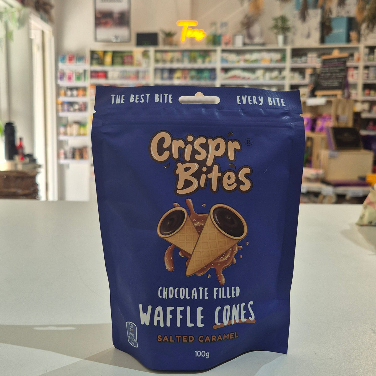 A blue stand-up pouch of Crispr Bites Chocolate Filled  Salted Caramel Waffle Cones on a white countertop in a shop setting.