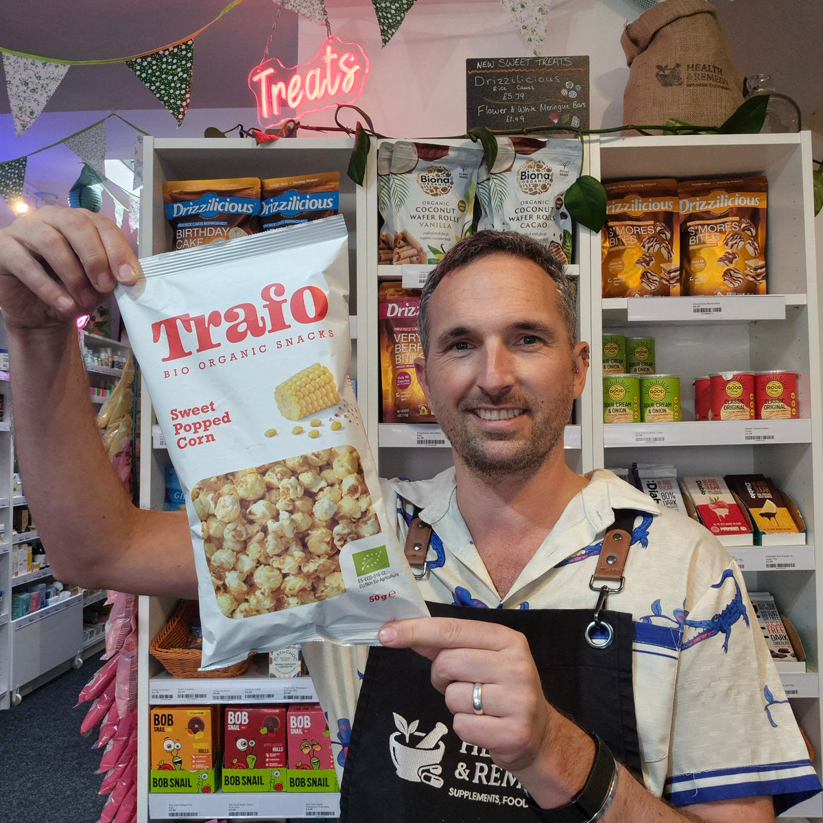 A man in a shop wearing an apron holds up a bag of Traifo Sweet Popped Corn, pointing at the front of the packaging while standing in front of shelves of snacks.