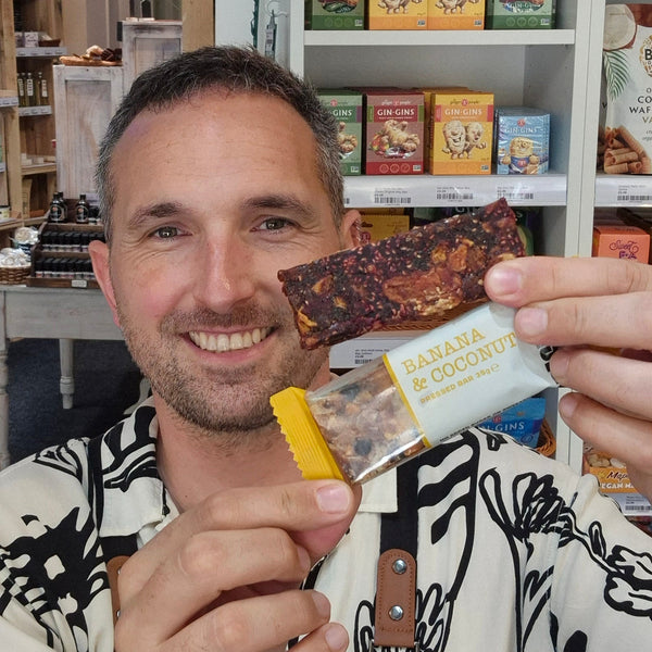 A smiling man in a patterned shirt and black Health & Remedy apron holds up two packaged Presteez snack bars in a shop setting with shelves and a pink neon 'Treats' sign in the background.
