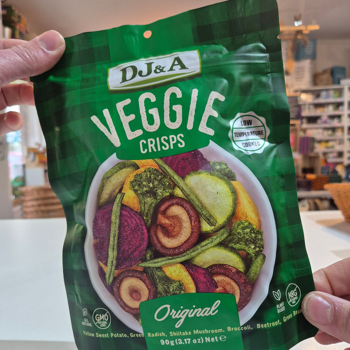 Hand holding a green package of DJ&A Veggie Crisps with colorful vegetable illustration on a blurred indoor background.