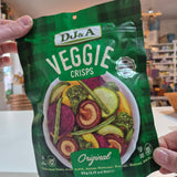 Hand holding a green package of DJ&A Veggie Crisps with colorful vegetable illustration on a blurred indoor background.