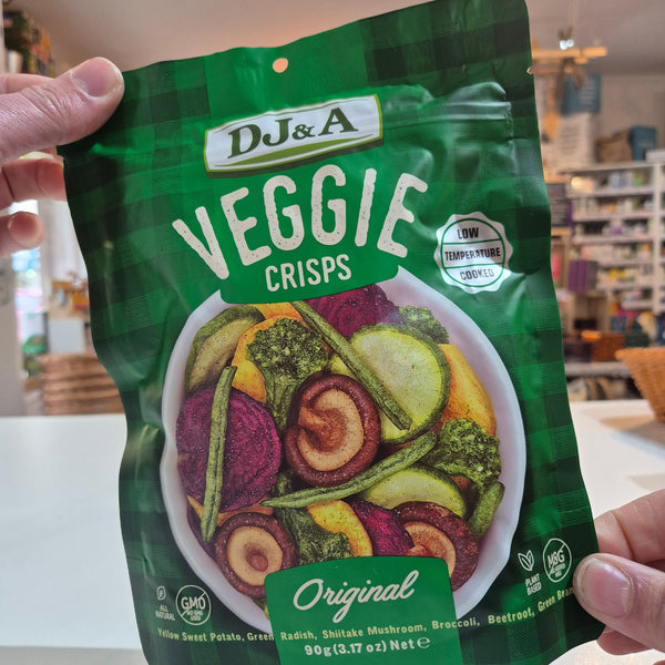 Hand holding a green package of DJ&A Veggie Crisps with colorful vegetable illustration on a blurred indoor background.