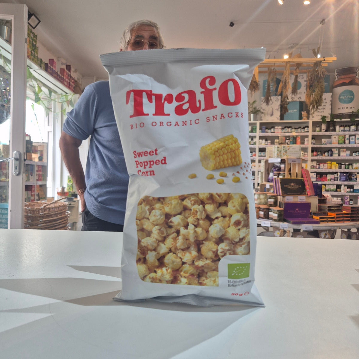 A bag of Trafo Sweet Popcorn organic snacks sits on a white counter in a retail store, with a man standing behind it.