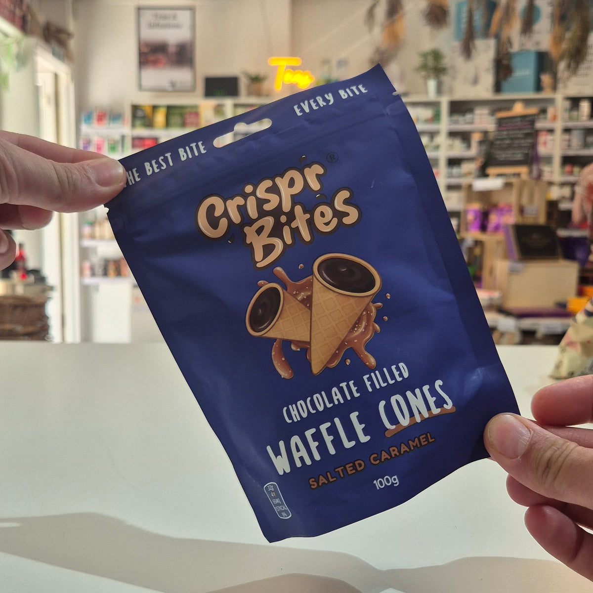 A person holds up a blue bag of Crispr Bites Chocolate Filled Waffle Cones in a shop setting.