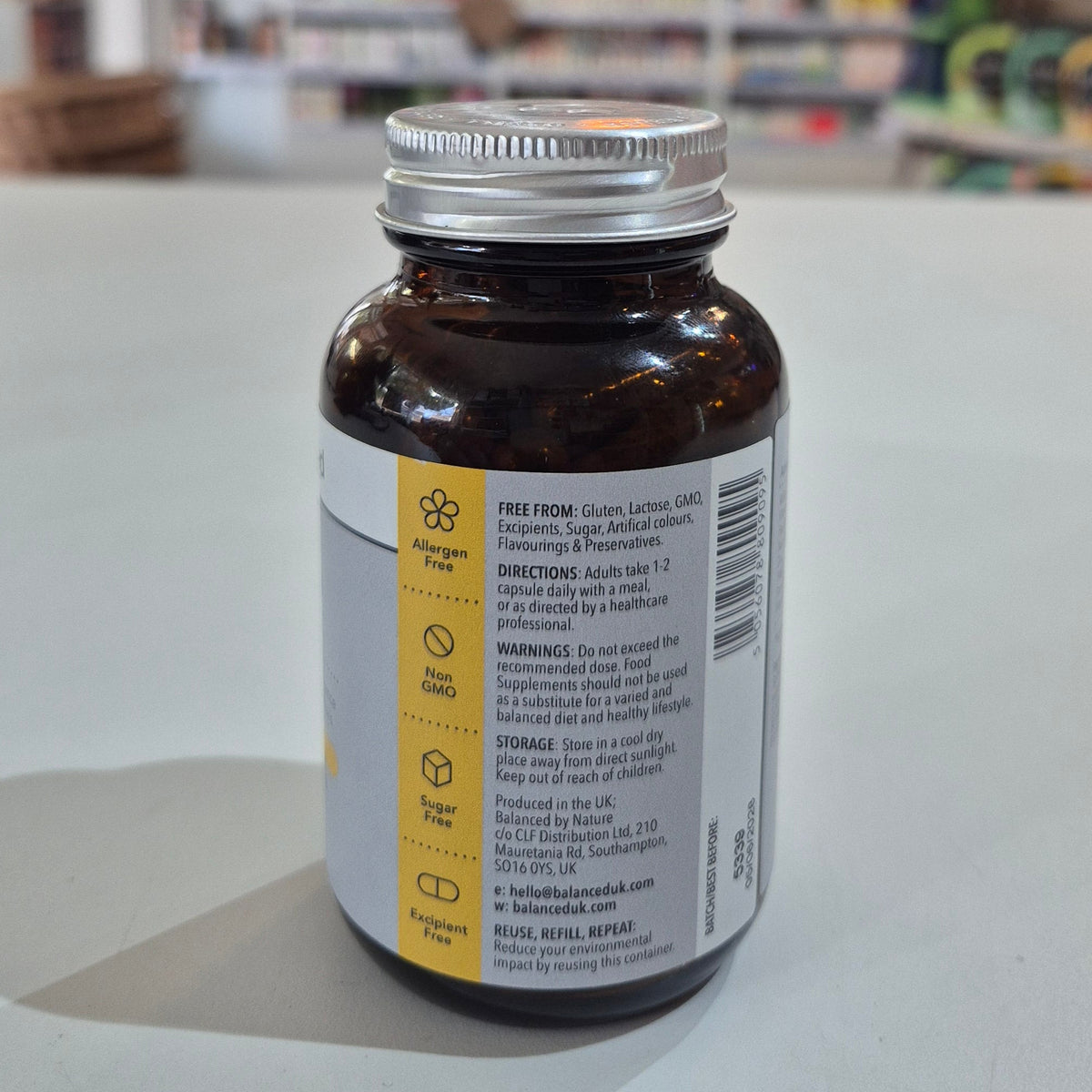 Supplement bottle labeled 'balanced' for hair, skin, and nails on a table with a blurred store background. Brown supplement bottle with label on a light gray surface