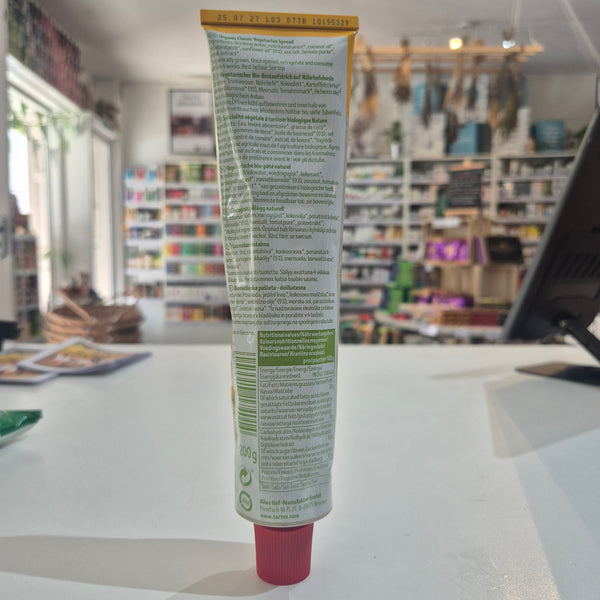 A tube of tartex classic vegetarian spread, with a blurred store interior in the background. The tube is mostly white with green text and a pink cap.