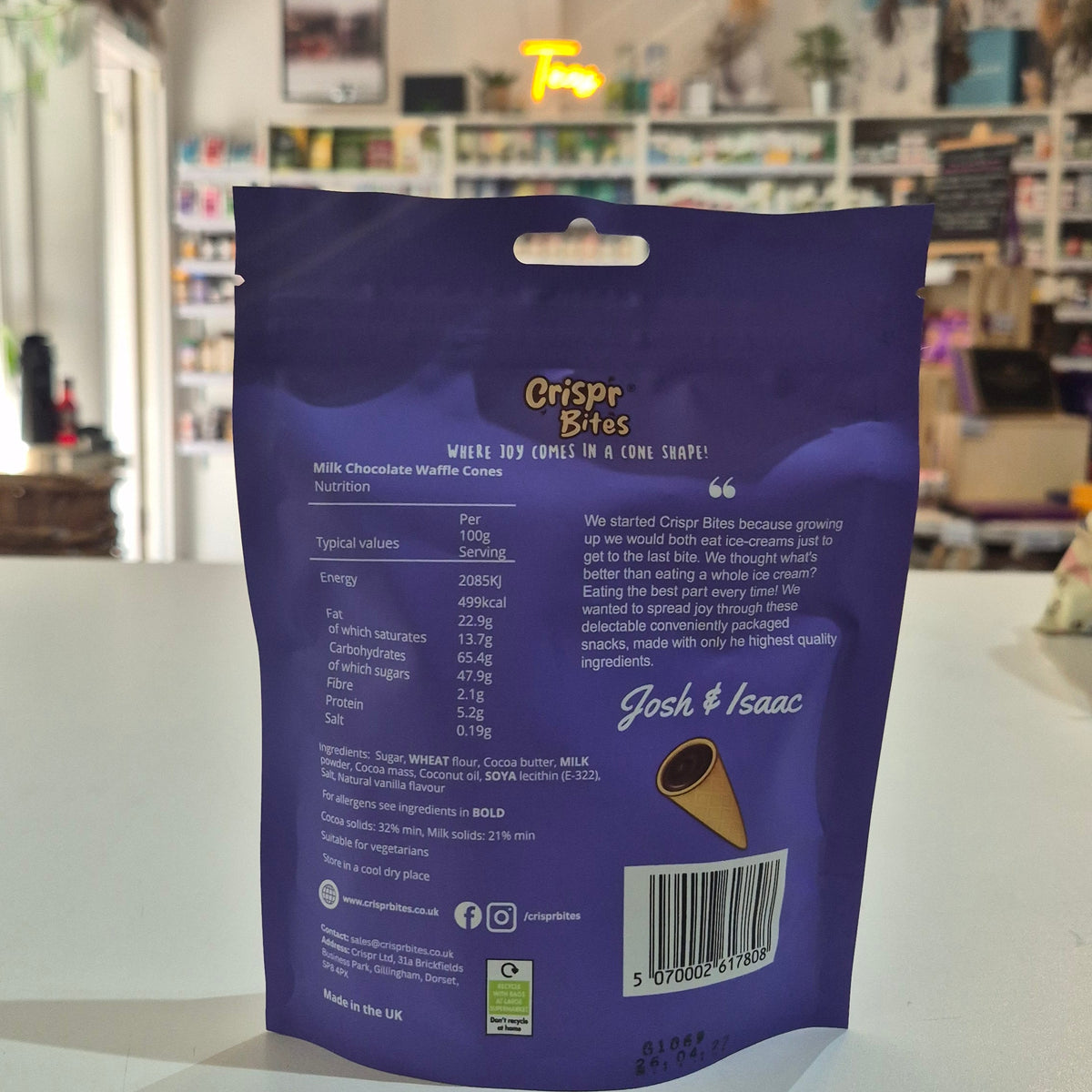 A purple stand-up pouch of Crispr Bites Milk Chocolate Filled Waffle Cones on a white countertop in a blurred retail shop setting with the ingredients showing.