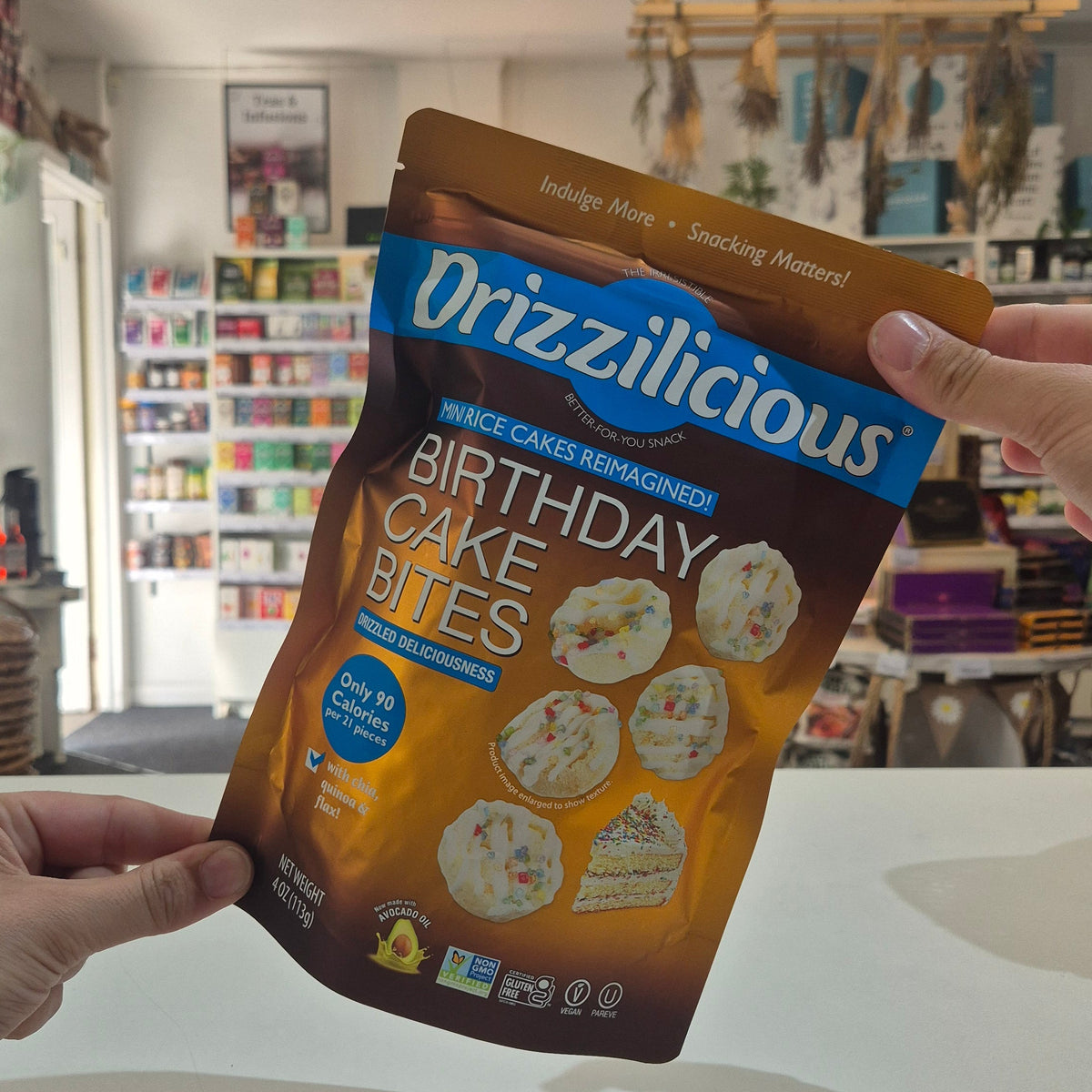 A person holds a bag of Drizzilicious Birthday Cake Bites in a retail store setting. The packaging is brown and blue, and the background shows store shelves and fixtures.