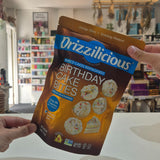 A person holds a bag of Drizzilicious Birthday Cake Bites in a retail store setting. The packaging is brown and blue, and the background shows store shelves and fixtures.