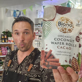 Man holding a package of Biona Organic Coconut Wafer Rolls Cacao in a store setting.