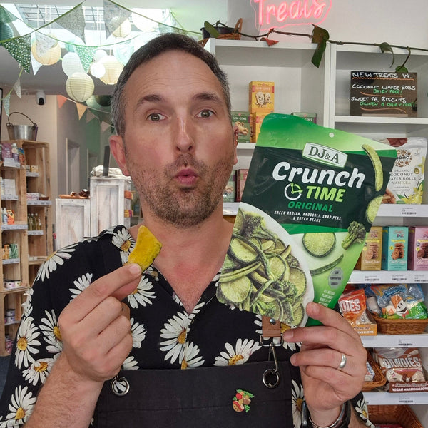 A man wearing a black apron with 'Health & Remedy' printed on it holds up a bag of DJ&A Crunch Time dog treats in one hand and a yellow treat in the other, standing inside a retail store.