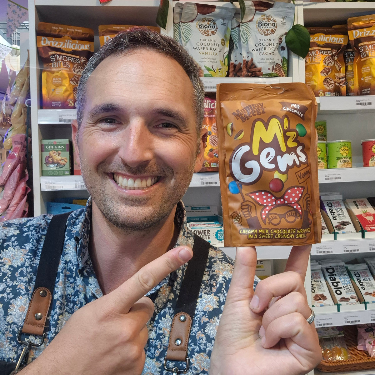 A smiling man in a patterned shirt and suspenders holds up a brown bag of Caramel M'z Gems chocolate gems in front of a store shelf filled with snacks.