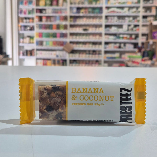 A single banana and coconut bar wrapper sits on a white tabletop in a blurred retail store setting. The wrapper is white with yellow side flaps and yellow text reading 'BANANA & COCONUT' and Presteez 'PRESSED BAR'.