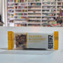 A single banana and coconut bar wrapper sits on a white tabletop in a blurred retail store setting. The wrapper is white with yellow side flaps and yellow text reading 'BANANA & COCONUT' and Presteez 'PRESSED BAR'.