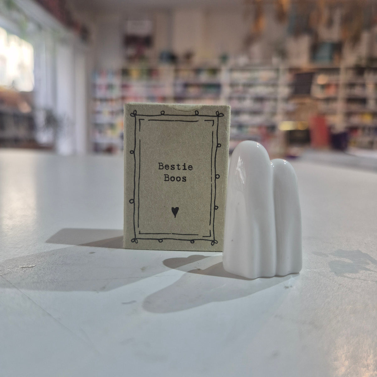 A small beige card labeled 'Bestie Boos' with a black border and heart, placed next to a white ceramic figurine shaped like three fingers, on a white tabletop in a blurred shop setting.
