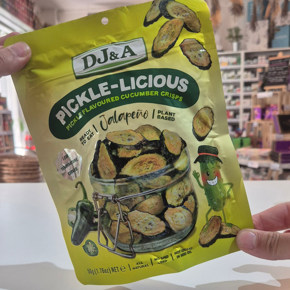 A person holds up a bag of DJ&A Pickle-Licious Jalapeño Pickled Gummy Cucumber Chips in a brightly lit store setting.
