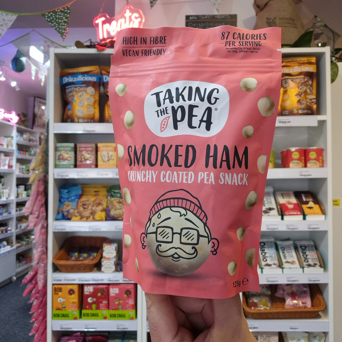 A hand holding a pink stand-up pouch of Taking the Pea Smoked Ham Crunchy Coated Pea Snack in a retail store setting with shelves of other snacks in the background.