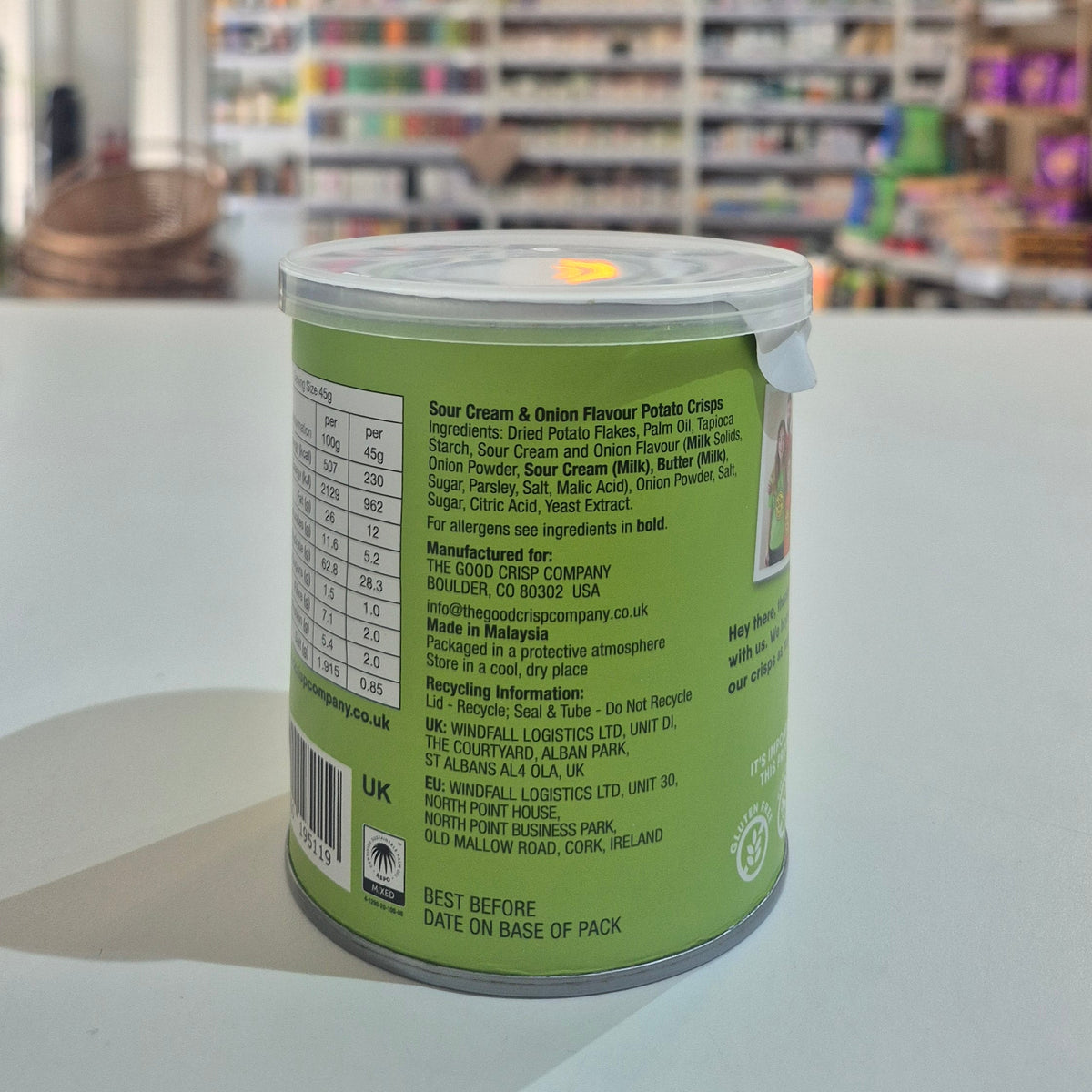 Can of 'Sour Cream & Onion' potato crisps from The Good Crisp Company on a white surface with a blurred store background. 