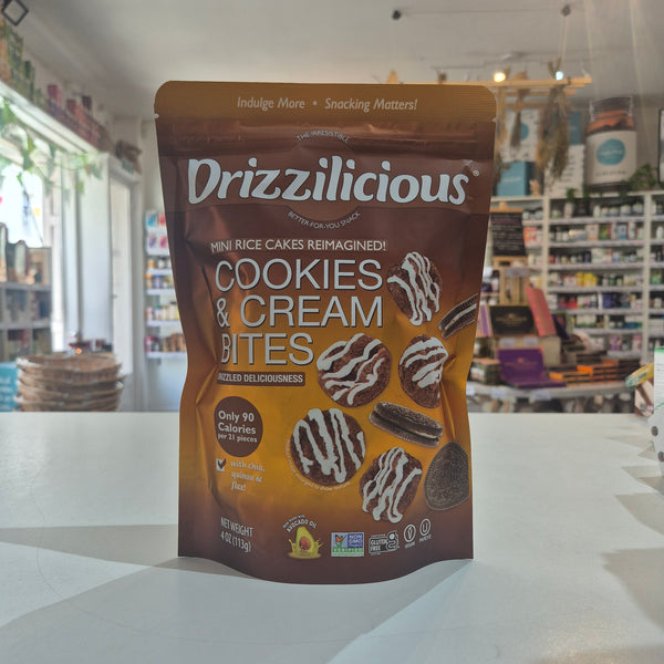 A stand-up pouch of Drizzilicious Cookies & Cream Bites mini rice cakes, displayed on a white countertop with a blurred bakery shop background.