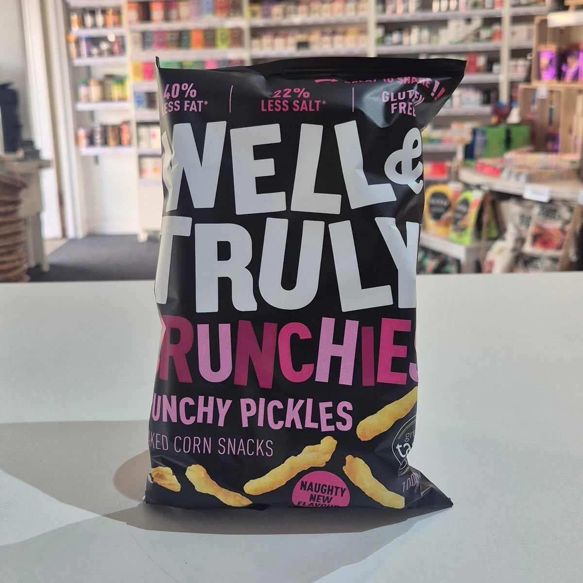 A bag of Well & Truly Crunchies Crunchy Pickles snack chips sits on a white counter in a retail store setting with shelves of products in the background.